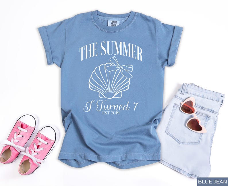 K&ouml;nnte beinhalten: Ein blaues T-Shirt mit dem Text "THE SUMMER I TURNED 7 EST 2019" und einer Muschel-Grafik. Das Shirt wird mit hellblauen Jeansshorts, einer rosa herzf&ouml;rmigen Sonnenbrille und rosa Turnschuhen kombiniert.