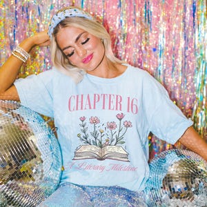 Puede incluir: Una camiseta azul claro con el texto "CHAPTER 16 A Literary Milestone" y un gr&aacute;fico de un libro abierto con flores que crecen de &eacute;l.