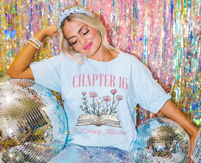 Puede incluir: Una camiseta azul claro con el texto "CHAPTER 16 A Literary Milestone" y un gr&aacute;fico de un libro abierto con flores que crecen de &eacute;l.