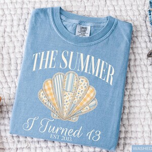May include: A light blue crew neck t-shirt featuring a graphic of a seashell composed of yellow gingham, blue striped, and floral patterned segments. The text reads THE SUMMER I Turned 13 EST 2013 in white lettering. The shirt has a Comfort Colors label.