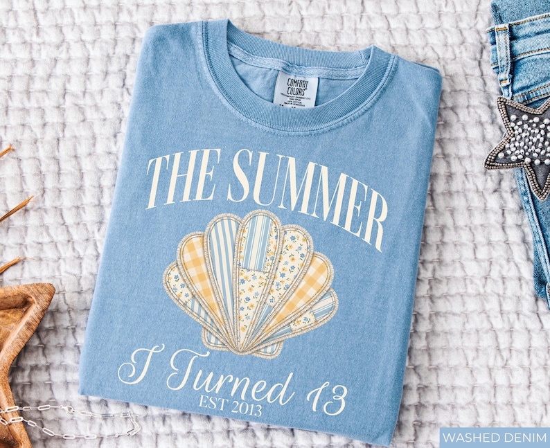 May include: A light blue crew neck t-shirt featuring a graphic of a seashell composed of yellow gingham, blue striped, and floral patterned segments. The text reads THE SUMMER I Turned 13 EST 2013 in white lettering. The shirt has a Comfort Colors label.