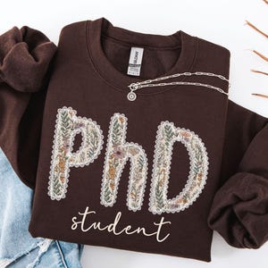 May include: A brown sweatshirt featuring the letters "PhD" decorated with a floral pattern, and the word "student" in script. A silver necklace with a pendant is draped across the sweatshirt. The sweatshirt is folded on top of blue jeans.