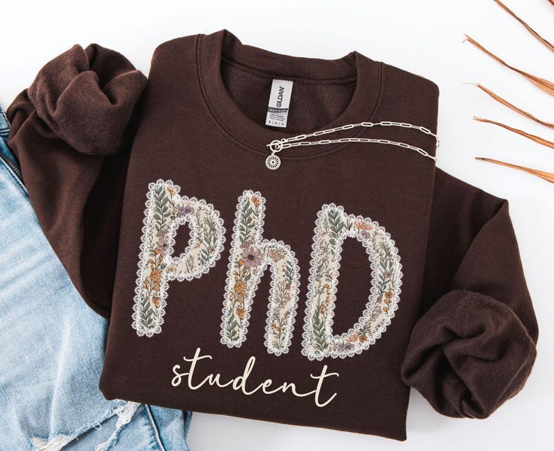 May include: A brown sweatshirt featuring the letters "PhD" decorated with a floral pattern, and the word "student" in script. A silver necklace with a pendant is draped across the sweatshirt. The sweatshirt is folded on top of blue jeans.