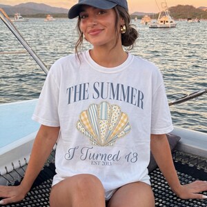 May include: A white short-sleeved t-shirt featuring a graphic of a seashell with blue, yellow, and white patterned segments. The text reads THE SUMMER I Turned 13 EST 2013 in blue lettering. The shirt has a crew neckline and a relaxed fit design.