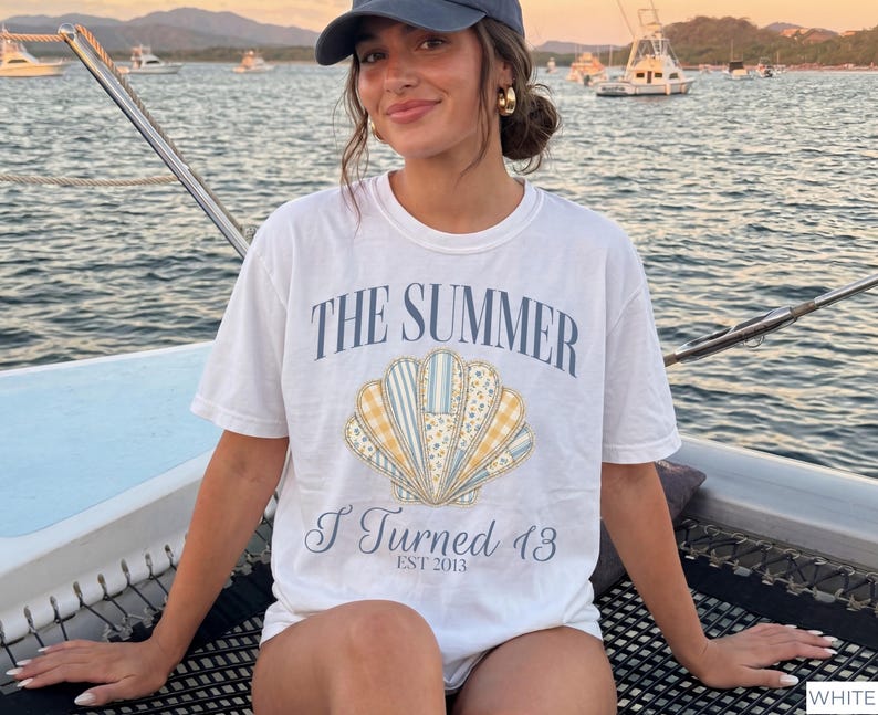 May include: A white short-sleeved t-shirt featuring a graphic of a seashell with blue, yellow, and white patterned segments. The text reads THE SUMMER I Turned 13 EST 2013 in blue lettering. The shirt has a crew neckline and a relaxed fit design.