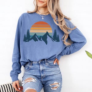 May include: A blue long-sleeved shirt with a graphic of a sunset over mountains and trees. The graphic is in a retro style with orange, yellow, blue, and green colors.