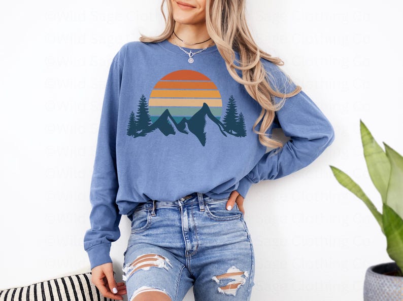 May include: A blue long-sleeved shirt with a graphic of a sunset over mountains and trees. The graphic is in a retro style with orange, yellow, blue, and green colors.