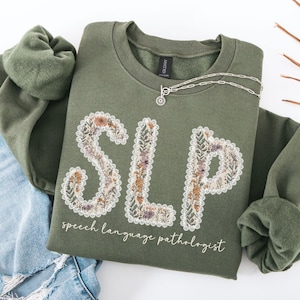 May include: An olive green sweatshirt with the letters "SLP" in a floral design. The words "speech language pathologist" are printed below the letters in a cursive font. A silver necklace is draped across the sweatshirt.
