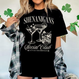 May include: Black t-shirt with white text that reads "Shenanigans Social Club Let's Get Shamrocked" and a graphic of three cocktails.