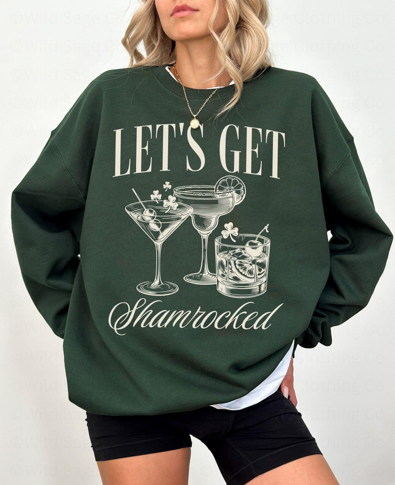 以下が含まれることがあります： 「Let's Get Shamrocked」という文字と、3つのカクテルのグラフィックが描かれた緑色のスウェットシャツ。