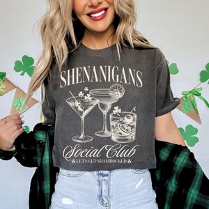 May include: A gray t-shirt with the text "Shenanigans Social Club" and a graphic of three cocktails. The text "Let's get shamrocked" is below the graphic.