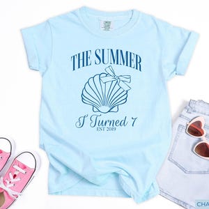 K&ouml;nnte beinhalten: Hellblaues T-Shirt mit dem Text "THE SUMMER I TURNED 7 EST 2019" und einer Muschel-Grafik. Das Shirt wird mit rosa Turnschuhen und Jeansshorts kombiniert, f&uuml;r einen l&auml;ssigen Sommer-Look.