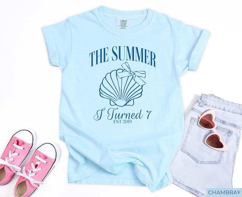 K&ouml;nnte beinhalten: Hellblaues T-Shirt mit dem Text "THE SUMMER I TURNED 7 EST 2019" und einer Muschel-Grafik. Das Shirt wird mit rosa Turnschuhen und Jeansshorts kombiniert, f&uuml;r einen l&auml;ssigen Sommer-Look.