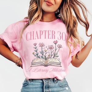 May include: Pink t-shirt with the text "CHAPTER 30 A Literary Milestone" and a graphic of an open book with flowers growing from it.