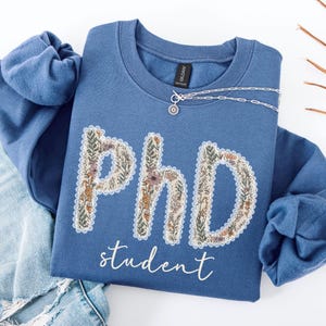 May include: A blue sweatshirt featuring the text "PhD student" in a floral design. The letters are filled with flowers and leaves. A silver necklace with a small pendant rests on the sweatshirt. The sweatshirt is folded on blue jeans.