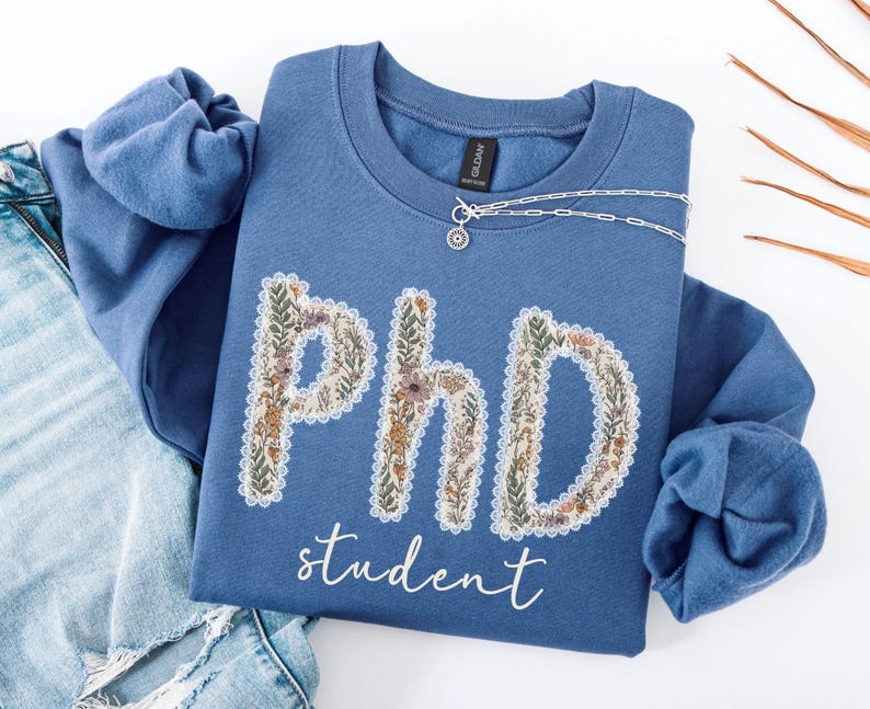 May include: A blue sweatshirt featuring the text "PhD student" in a floral design. The letters are filled with flowers and leaves. A silver necklace with a small pendant rests on the sweatshirt. The sweatshirt is folded on blue jeans.