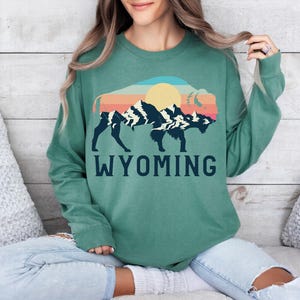 May include: A green sweatshirt with a vintage-style graphic of a bison silhouette with mountains and a sun in the background. The text "WYOMING" is printed below the graphic.