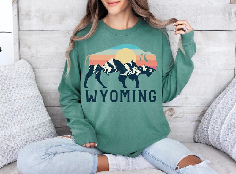 May include: A green sweatshirt with a vintage-style graphic of a bison silhouette with mountains and a sun in the background. The text "WYOMING" is printed below the graphic.