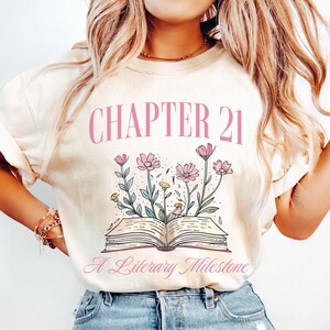 May include: A white t-shirt with the text "Chapter 21 A Literary Milestone" in pink lettering. The design features a book with flowers around it.