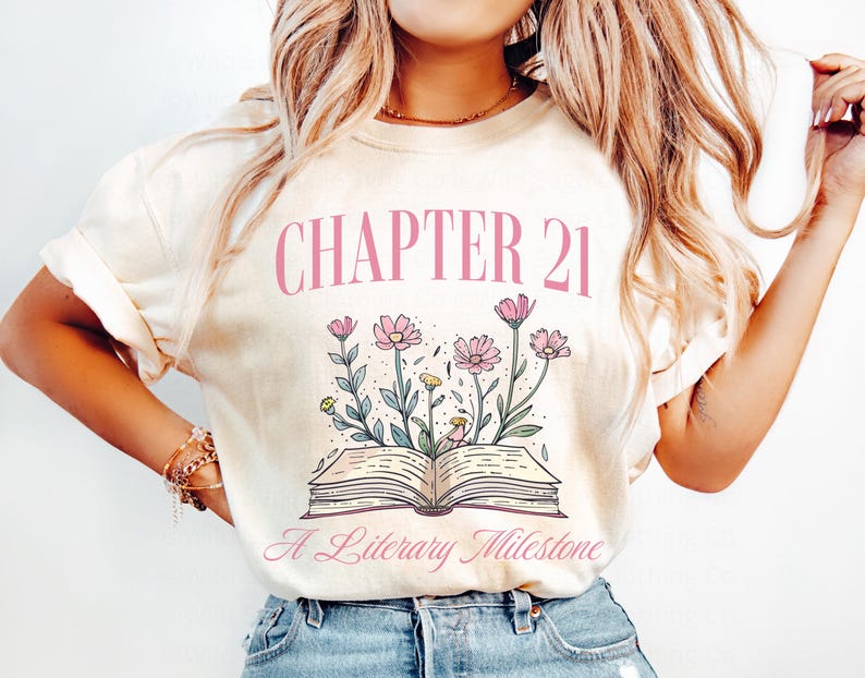 May include: A white t-shirt with the text "Chapter 21 A Literary Milestone" in pink lettering. The design features a book with flowers around it.