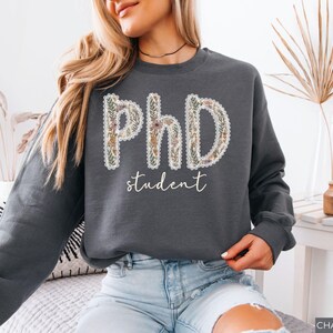 May include: Dark grey sweatshirt featuring the text "PhD student". The letters "PhD" are decorated with a floral pattern, and the word "student" is written in a cursive font. The sweatshirt is a relaxed fit and a casual style.