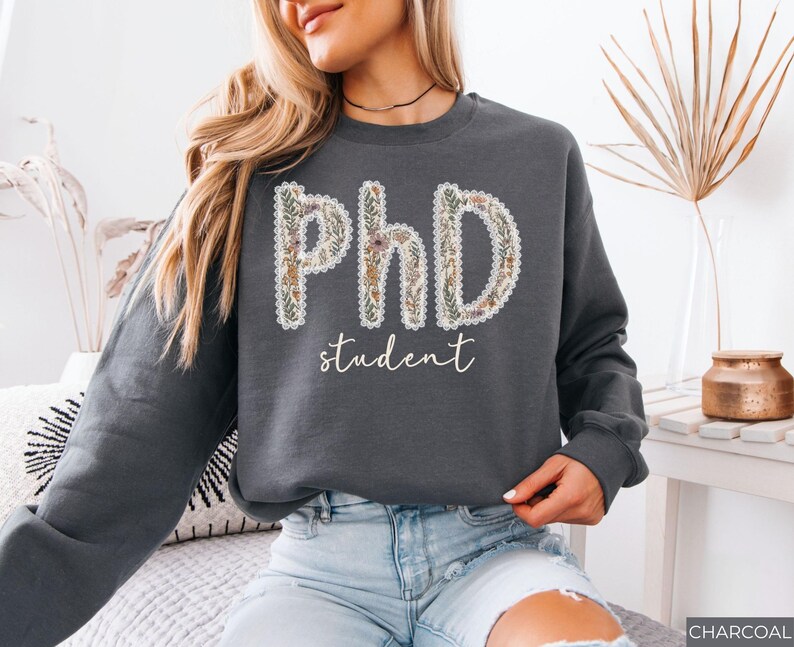 May include: Dark grey sweatshirt featuring the text "PhD student". The letters "PhD" are decorated with a floral pattern, and the word "student" is written in a cursive font. The sweatshirt is a relaxed fit and a casual style.