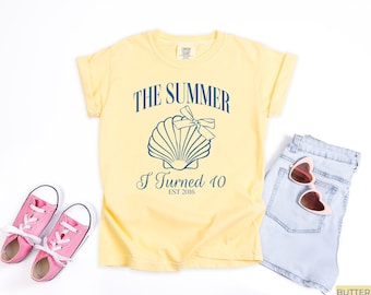 Puede incluir: Camiseta amarilla clara con el texto "THE SUMMER I TURNED 10 EST 2016" y un diseño de concha. La camiseta se combina con pantalones cortos vaqueros azul claro, gafas de sol rosas y zapatillas rosas.