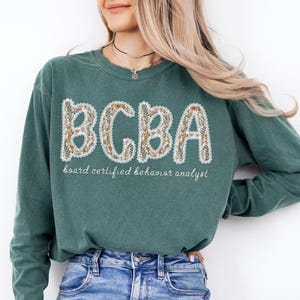 May include: A teal green sweatshirt with the letters "BCBA" in a floral design. Below the letters, the text reads "board certified behavior analyst" in a script font. The sweatshirt is a crew neck style.