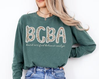 May include: A teal green sweatshirt with the letters "BCBA" in a floral design. Below the letters, the text reads "board certified behavior analyst" in a script font. The sweatshirt is a crew neck style.