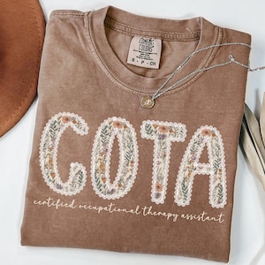 Può includere: Una t-shirt marrone a maniche corte con scollo rotondo. La parte anteriore presenta la scritta COTA in grandi lettere riempite con un motivo floreale e bordate di pizzo bianco, con sotto il testo certified occupational therapy assistant.