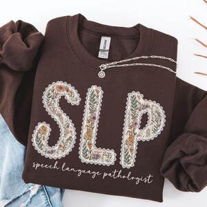 May include: A brown sweatshirt with the letters "SLP" in a floral design. The words "speech language pathologist" are written below the letters in a script font. A silver necklace is draped across the sweatshirt.