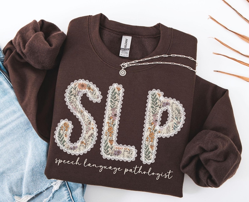 May include: A brown sweatshirt with the letters "SLP" in a floral design. The words "speech language pathologist" are written below the letters in a script font. A silver necklace is draped across the sweatshirt.
