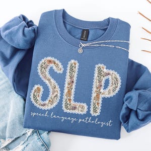 May include: A blue crew neck sweatshirt with the letters "SLP" in a floral design. The words "speech language pathologist" are written below the letters in a script font. A silver necklace is draped across the sweatshirt.
