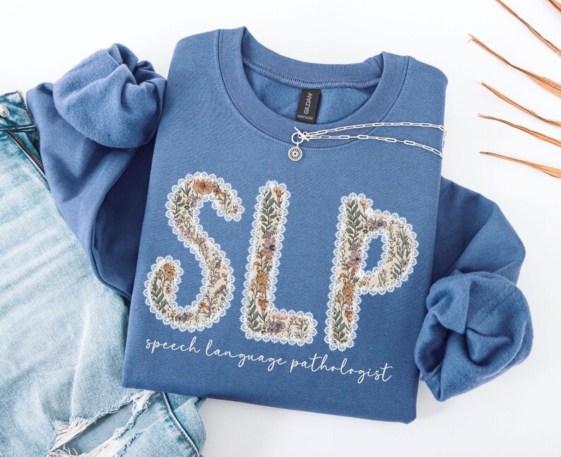 May include: A blue crew neck sweatshirt with the letters "SLP" in a floral design. The words "speech language pathologist" are written below the letters in a script font. A silver necklace is draped across the sweatshirt.