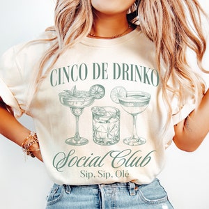 May include: A beige t-shirt with the text "Cinco De Drinko Social Club Sip, Sip, Ole" in green lettering. The shirt features a graphic of three cocktails: a margarita, a martini, and an old fashioned.