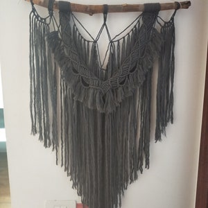 May include: A gray macrame wall hanging with a wooden branch at the top. The macrame is made with a thick cord and has a fringe at the bottom.