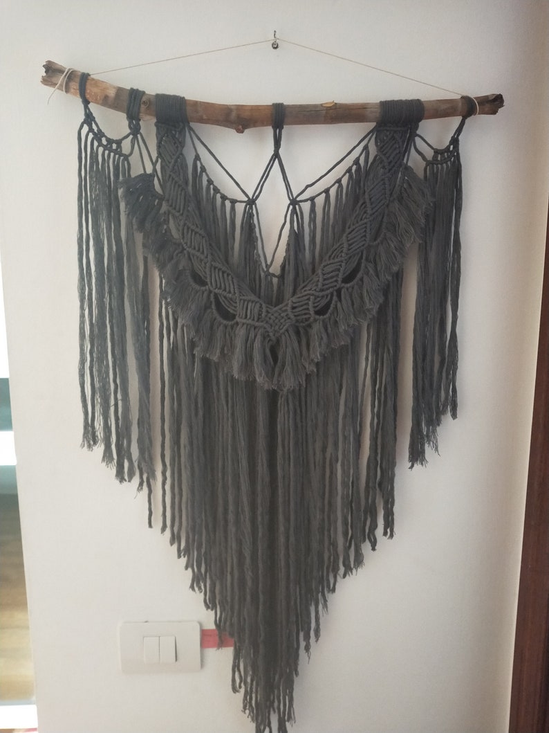 May include: A gray macrame wall hanging with a wooden branch at the top. The macrame is made with a thick cord and has a fringe at the bottom.