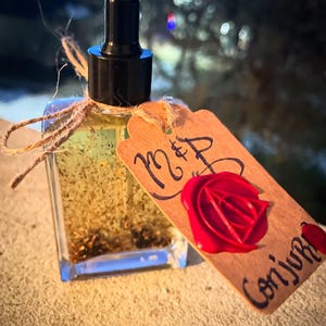 May include: A small glass bottle with a dropper top filled with a clear liquid and gold glitter. The bottle has a brown paper tag with the text "M&P" and a red wax seal with a triangle symbol. The tag also has the text "Conjuro" written on it.