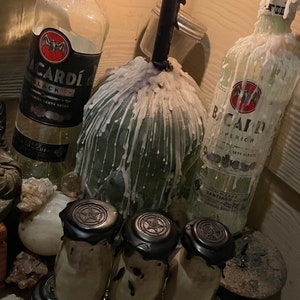 May include: A collection of six small glass bottles with black lids and a pentagram design. The bottles are arranged in a pyramid shape and are surrounded by white wax drippings. Two bottles of Bacardi rum are visible in the background, one with a black label and the other with a white label. A lit candle is also visible in the background.