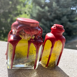 May include: Two glass bottles with red wax drippings, filled with a yellow liquid and black seeds. The bottles are on a gray surface.