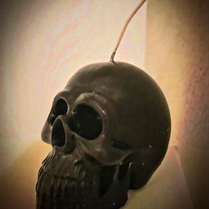 May include: A black skull-shaped candle with a wick. The candle has detailed facial features, including eye sockets, a nose cavity, and a jaw. The candle is a dark, matte black color, and the wick is a light brown.