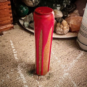 May include: A cylindrical glass container with a red wax top and red wax drips down the sides. The interior appears to be filled with a light brown material. The container is standing on a textured surface.