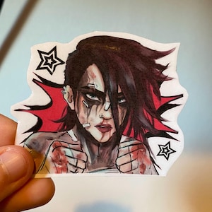 以下が含まれることがあります： 黒い髪に包帯と白いシャツを着た女性の漫画イラストが描かれたステッカー。彼女は獰猛な表情で拳を突き上げている。背景は赤で、黒い星が散りばめられている。