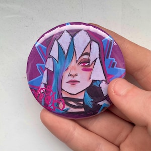 May include: A circular badge with a purple base, displaying a cartoon illustration of a person with blue and white hair, pink eye makeup, and a black choker. The design includes blue and pink accents, creating a stylized look.