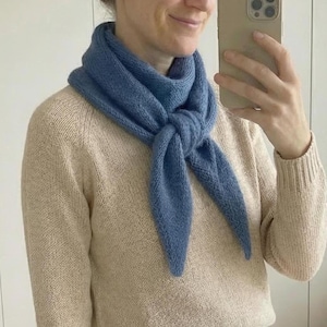 May include: A blue knitted scarf tied in a knot, worn with a beige jumper and white trousers. A wicker basket containing dark clothes is visible in the background.