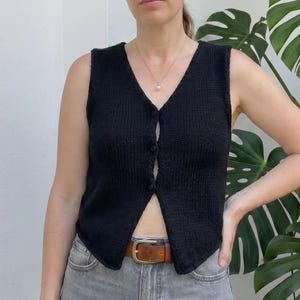 May include: A black, sleeveless, button-down vest with a V-neck. The vest is paired with gray jeans and a brown belt. The background is a white wall and a large green plant.