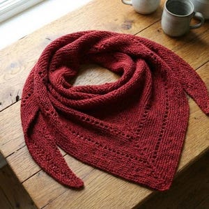 May include: A knitted triangular shawl in a deep red hue, displayed on a wooden surface. The shawl features a textured stitch pattern and a decorative border with small openings. The cozy accessory is folded to showcase its shape.