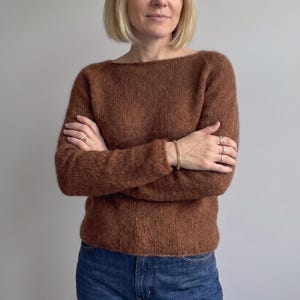 May include: A long-sleeved, brown knit sweater featuring a wide boat neckline and a fuzzy, textured surface. The garment is paired with blue denim jeans, creating a simple and functional outfit with a consistent, solid earth-tone color palette.