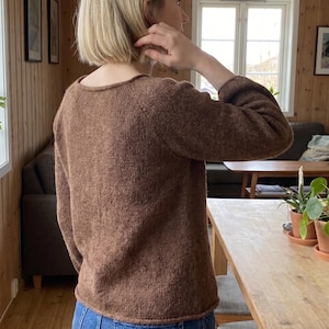 May include: A long-sleeved, brown knit sweater featuring a round neckline and a textured, heathered appearance. The garment has a simple, straight-cut silhouette with ribbed detailing along the hem and cuffs, providing a consistent, solid color.