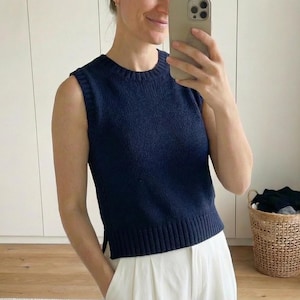 May include: A navy blue sleeveless jumper with a ribbed hem and crew neck. The jumper is worn with white trousers. A wicker basket filled with clothes is in the background.